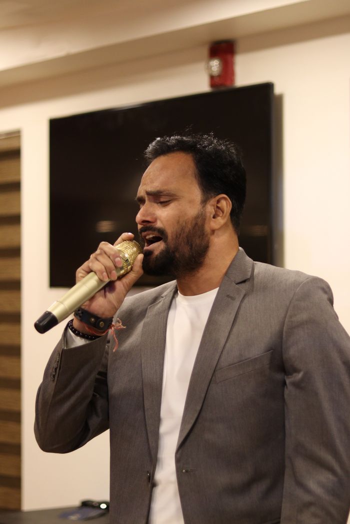 about-img A man in a suit sings passionately into a microphone at an indoor event.
